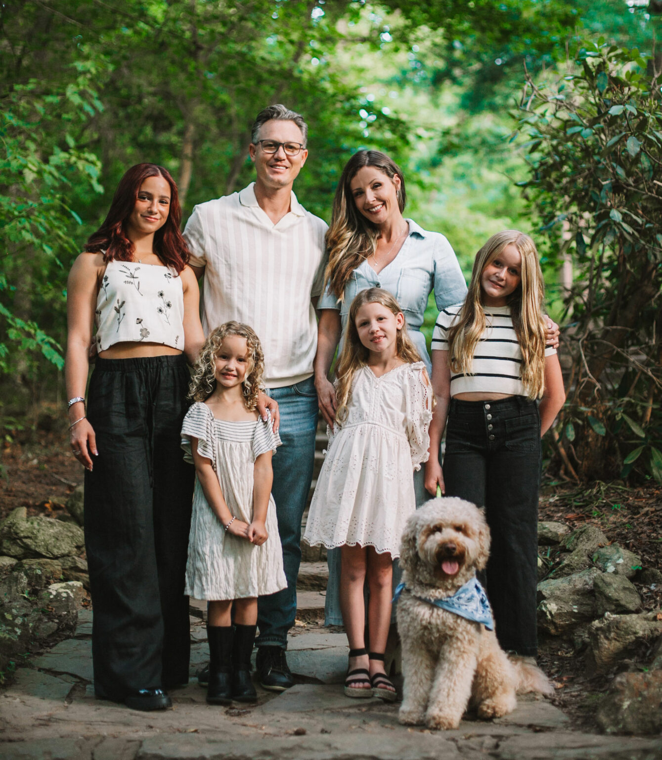 A family of six, including two adults, three young girls, a teenage girl, and a fluffy dog with a bandana, pose together on a stone path in their lush yard—the perfect backdrop for their recent home renovation in Charlotte.
