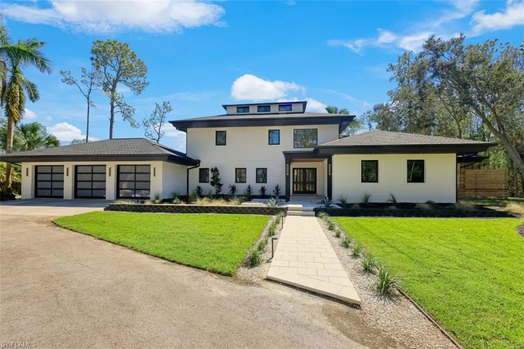Modern two-story house with a white exterior, dark trim, and large windows. Updated by a top remodeling contractor Charlotte NC, it features a three-car garage on the left, landscaped yard, and is surrounded by trees under a blue sky.
