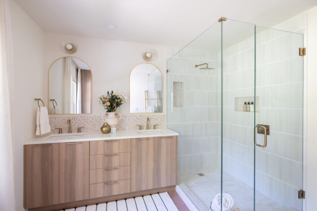Modern bathroom with a glass-enclosed shower, gold fixtures, dual sinks with wood cabinetry, two arched mirrors, a flower vase, and a white striped rug on the floor.