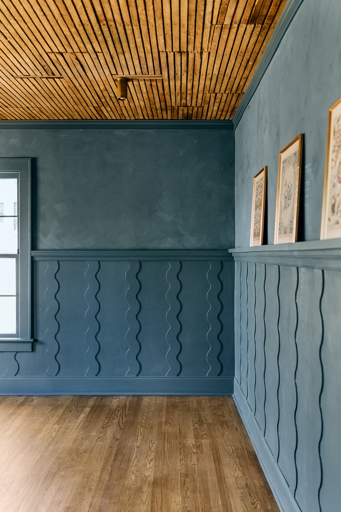 A room with blue textured walls featuring wavy patterns, a wooden slatted ceiling, hardwood floors, a window on the left, and three framed pictures on the wall to the right.