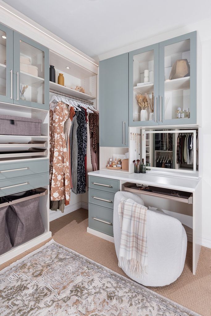 A modern, organized walk-in closet with light blue cabinets, hanging clothes, shelves with accessories, a built-in vanity with a mirror, a white chair draped with a plaid blanket, and a patterned rug on the floor.