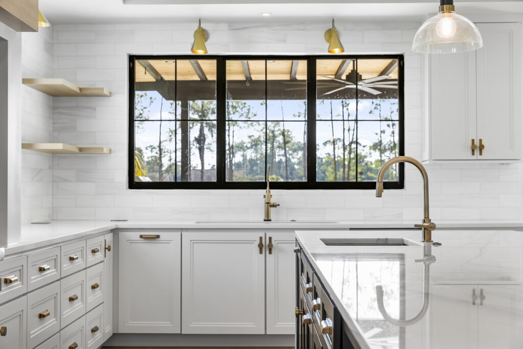 Bright modern kitchen with white cabinets, gold hardware, marble countertops, large window above the sink, open wooden shelves, and pendant lights, creating a clean and spacious look.