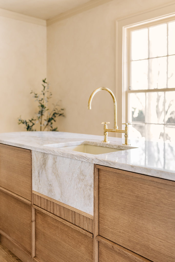 A modern kitchen with a marble farmhouse sink, gold faucet, light wood cabinets, and a large window letting in natural light. A plant is in the background.