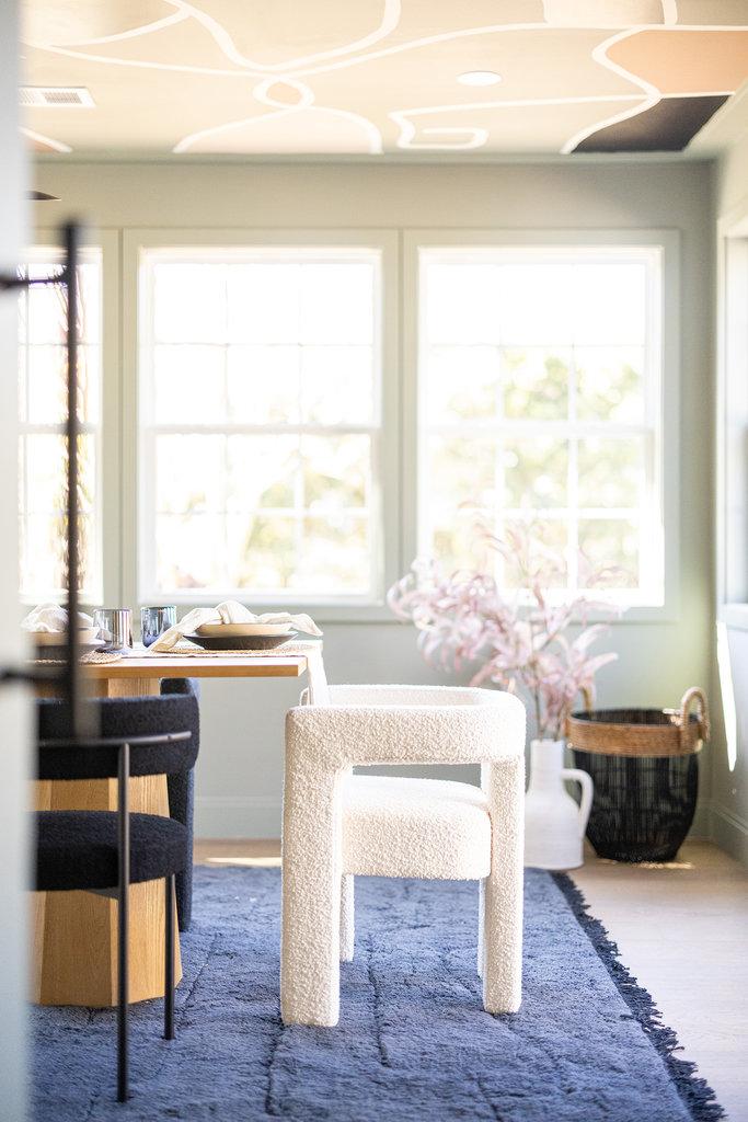 A bright, modern dining room with large windows, a light wooden table set for a meal, boucle white and black chairs, a blue rug, and a vase holding pink artificial branches near a black basket.